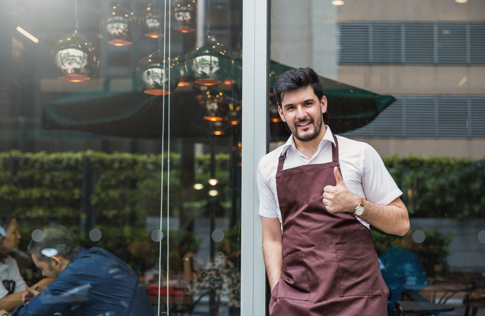 Dueño de negocio latino frente a su restaurante con el pulgar hacia arriba para ilustrar la idea de "construir tu negocio"