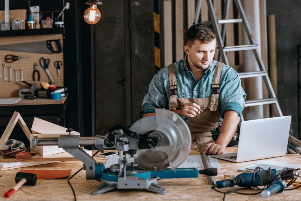Young bearded carpenter focused on planning woodworking project using laptop in workshop. Concept: best laptops for business