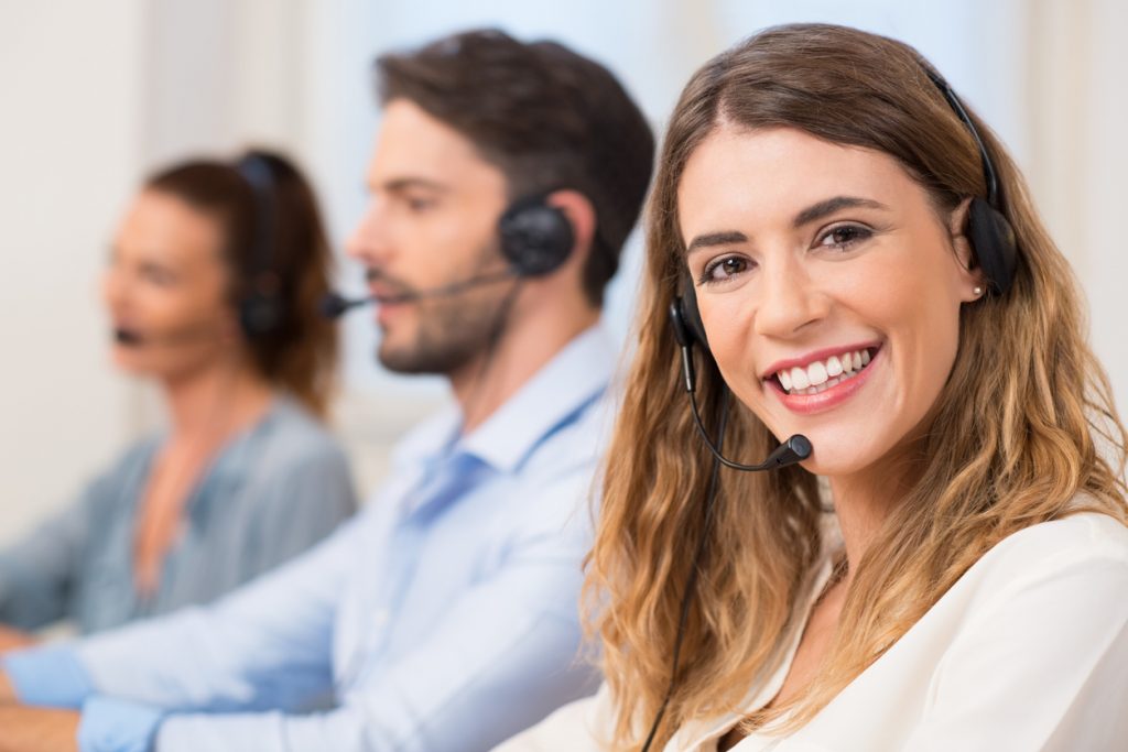 Smiling female call centre operator doing her job with a headset while looking at the camera. Portrait of happy woman in a call center smiling and working. Portrait of happy smiling female customer support phone operator at workplace. Concept: customer database