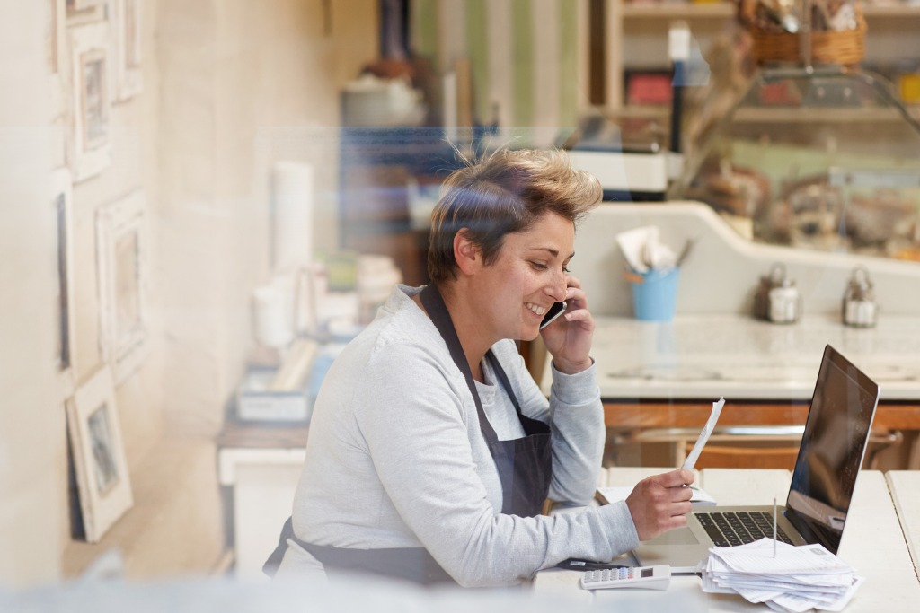 Female business owner with apron on computer and making calls negotiating her high interet rates on a business loan.