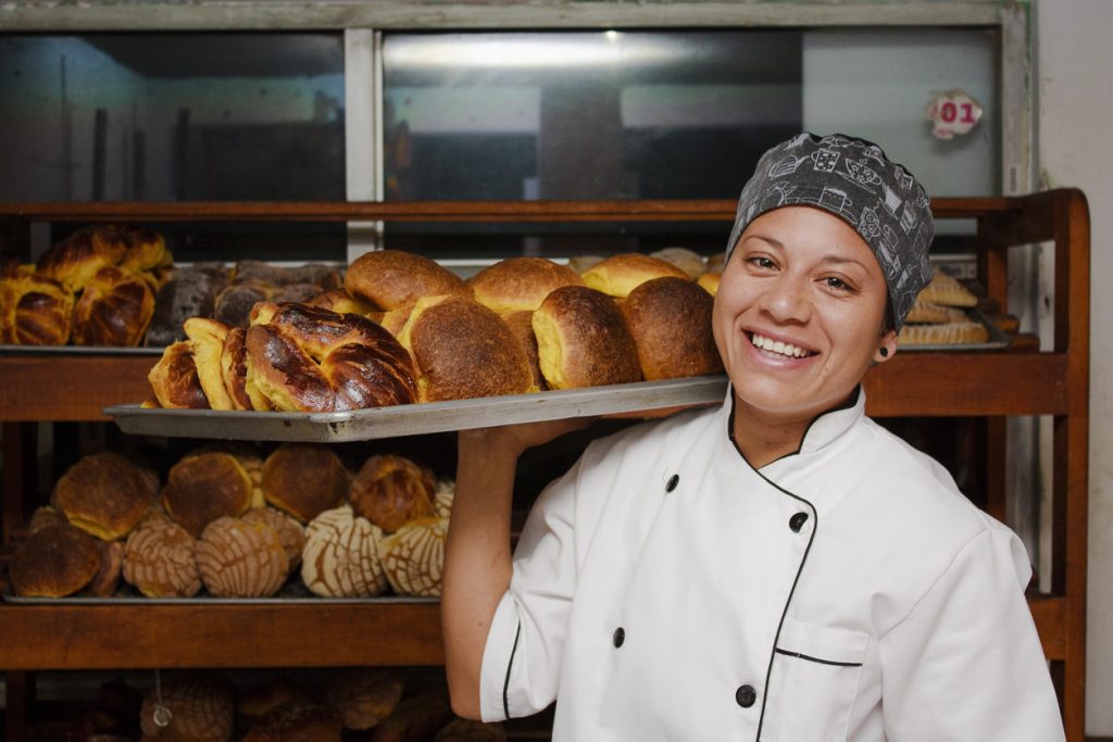 Empresaria inmigrantes: dueña de panadería sonriente cargando una bandeja de productos recién horneados. Concepto: préstamos para inmigrantes.