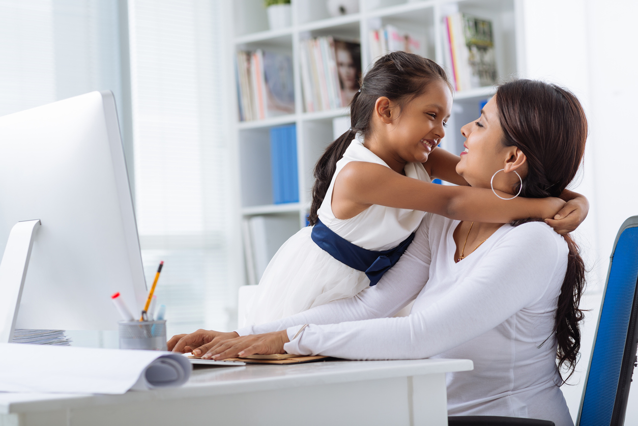 Young daughter embracing her mompreneur while she sitting at office desk and working on project