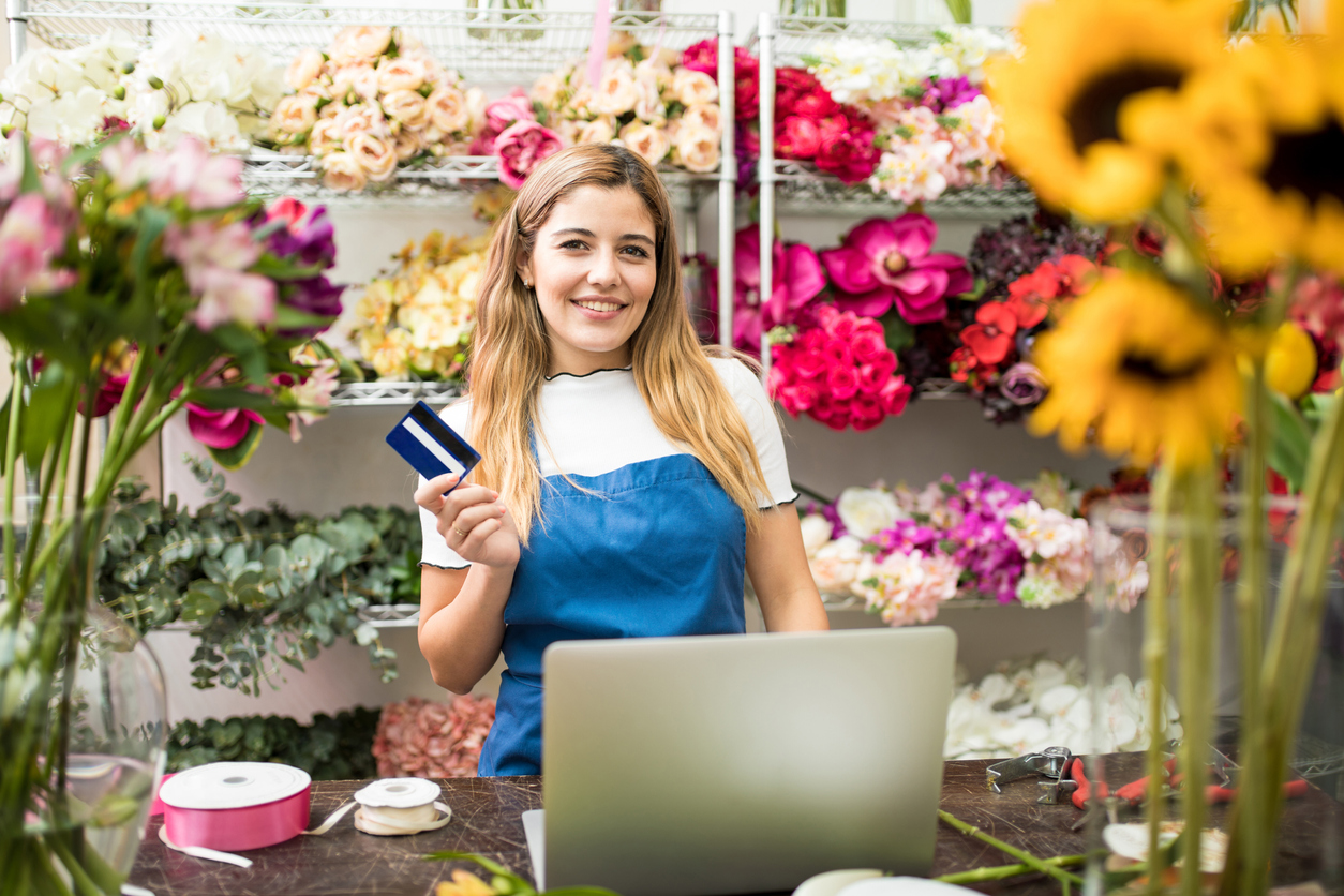 Flower shop owner holding a credit card next to a laptop computer in her flower shop and smiling. Concept: zero interest credit cards