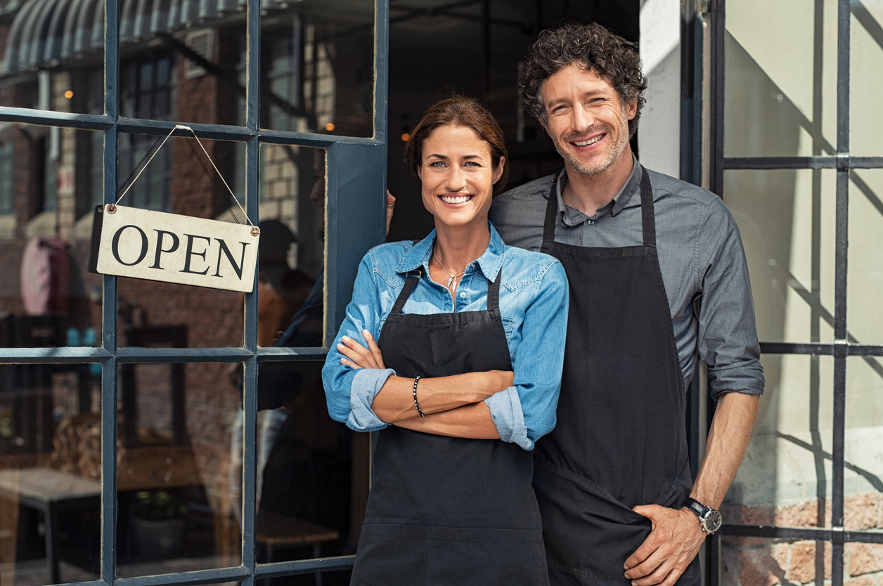Couple of small business owners smiling and looking at camera while standing at business entrance door with open sign board. Concept: business license
