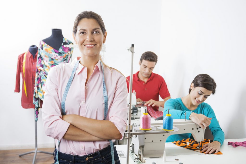 Portrait of smiling tailor with arms crossed standing at sewing factory with employees working in background. Concept: What's the difference between registering and incorporating a business?