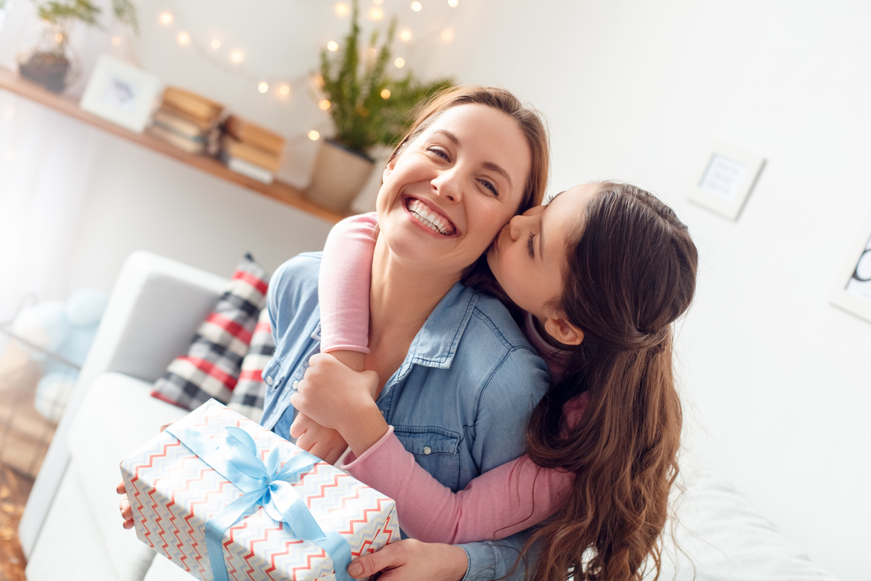 Young woman and girl at home celebrating mother's day sitting on sofa daughter hugging mother kissing cheek mom laughing joyful holding gift box. Concept: Mother's Day ideas