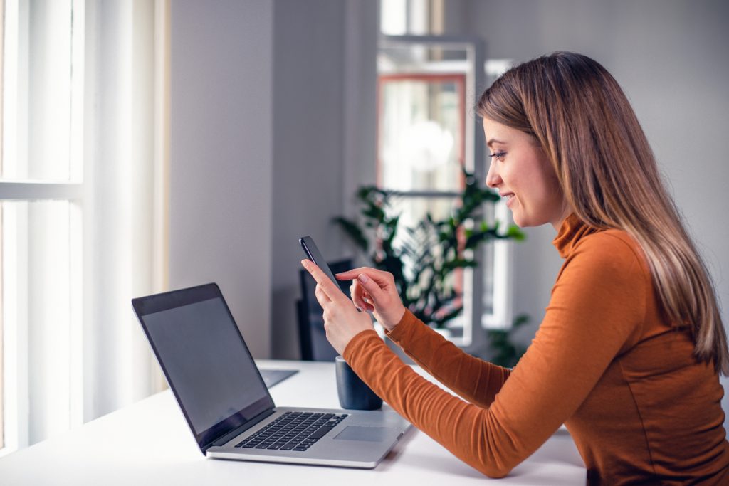 Woman using phone app while working at the office in front of the laptop. Browsing internet, doing online shopping, making reservations. Concept: how to pay off a loan