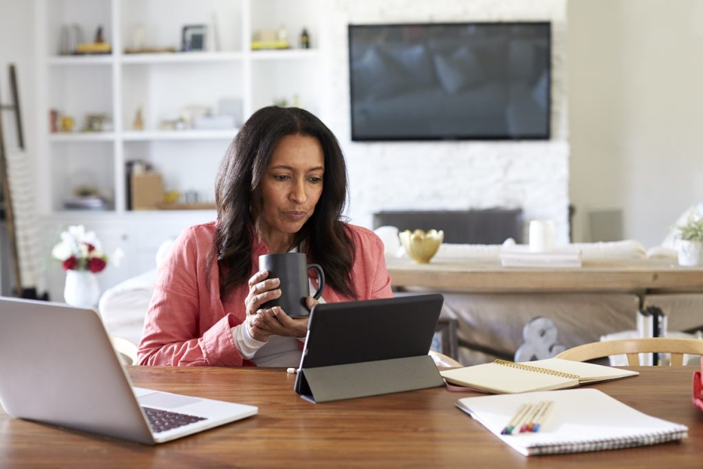 Middle aged woman sitting at a table reading using a tablet computer, holding a cup, front view. Concept: Apply for a business loan