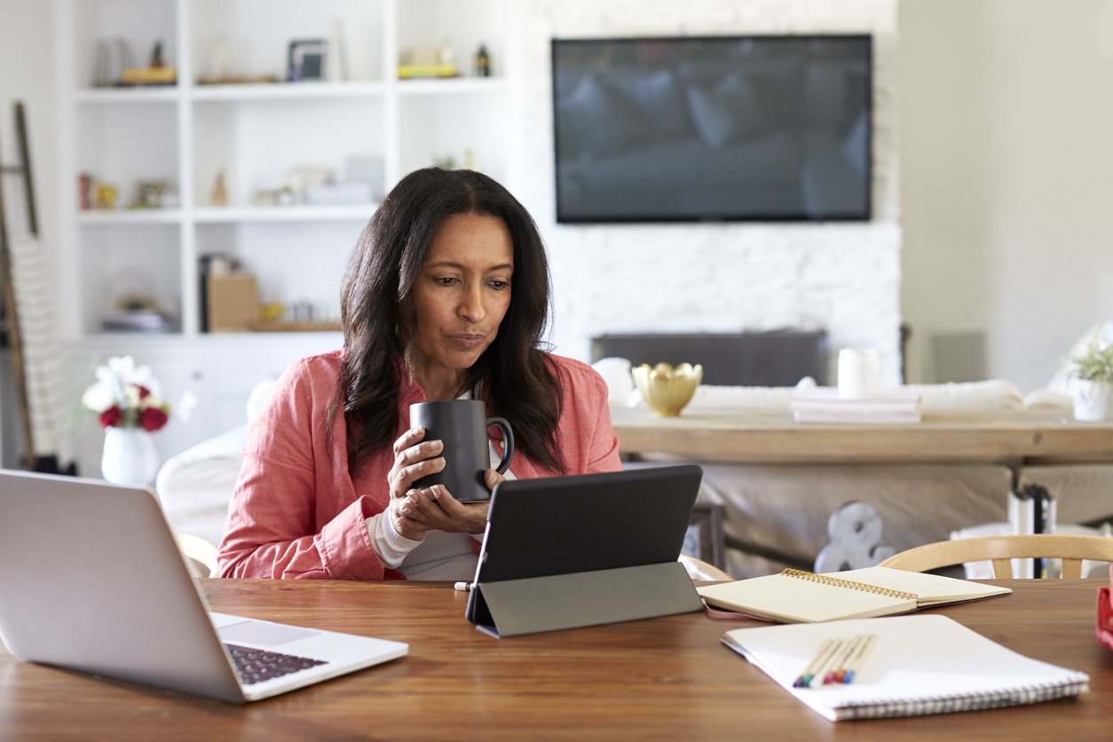 Middle aged woman sitting at a table reading using a tablet computer, holding a cup, front view. Concept: Apply for a business loan