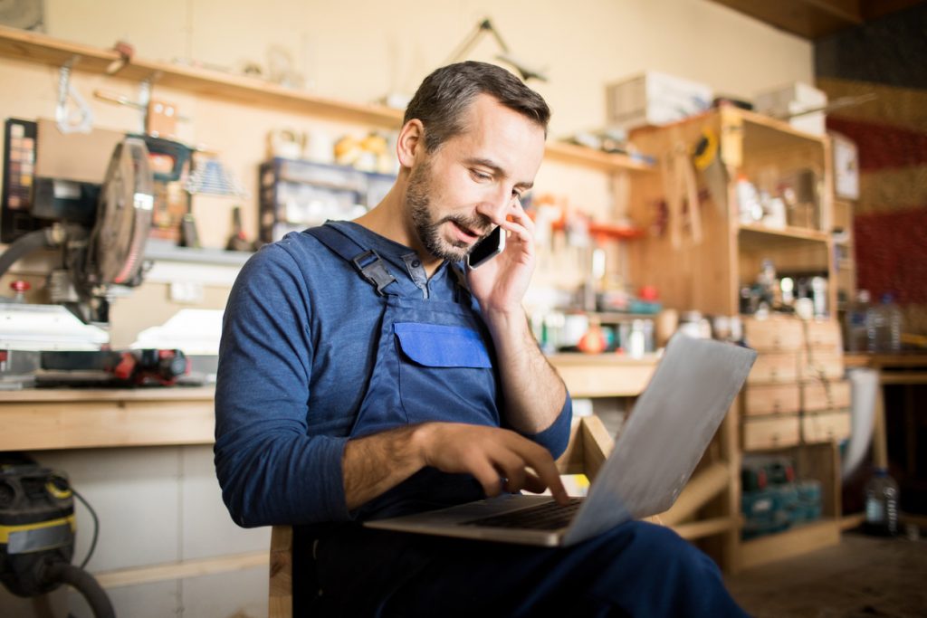Small business owner using laptop and speaking by smartphone in workshop. Concept: business savings account.