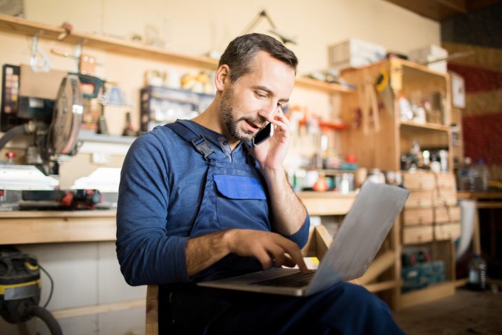 Small business owner using laptop and speaking by smartphone in workshop. Concept: business savings account.