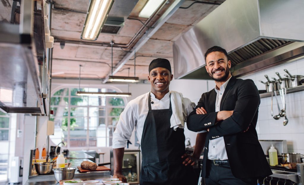 Portrait of restaurant owner with chef in kitchen. Businessman with professional cook standing together and looking at camera. Concept: restaurant revenue