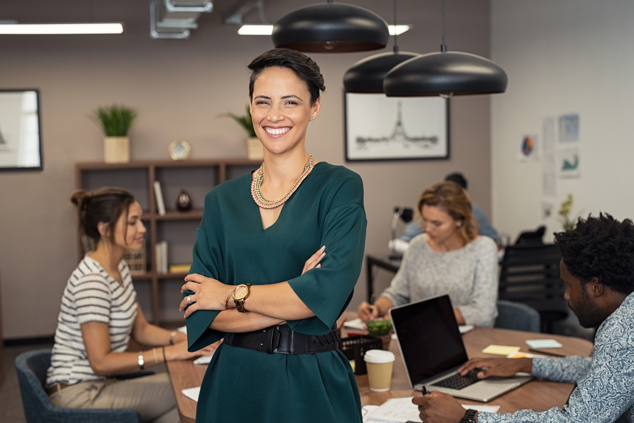 Portrait of successful business woman standing with her colleagues working in background at office. Portrait of cheerful fashion girl in green dress standing with folded arms and looking at camera. Beautiful businesswoman feeling proud and smiling. Concept: micrloans