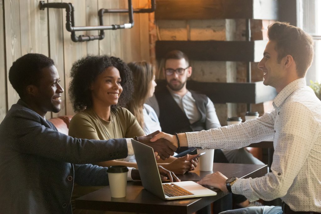 Multiracial african and caucasian men handshaking at meeting in cafe, diverse friends greeting sitting at coffeeshop table or black couple making deal shaking hands to white businessman with laptop. concept: Microloans vs the 3 F's