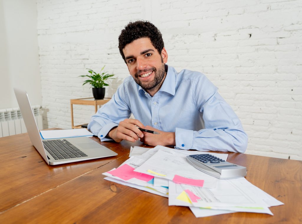 Happy attractive entrepreneur man calculating costs, charges, mortgage, taxes or paying bills with documents and laptop at home office. In online banking and Success business finances free of debts.