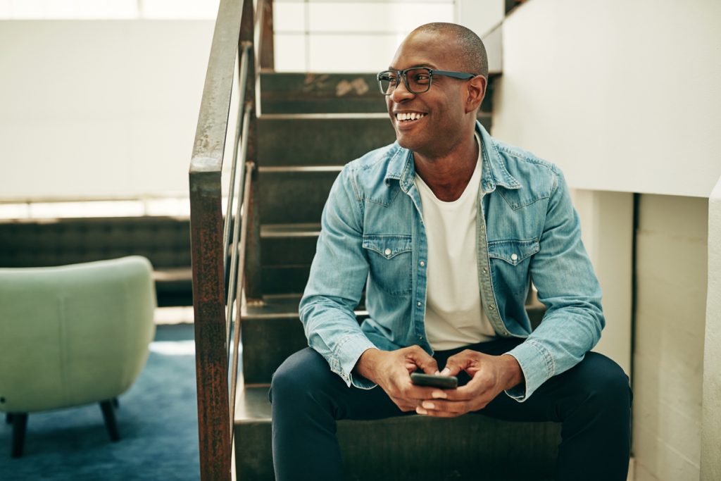 Empresario joven sonriendo, tiene gafas/lentes y un celular en la mano. Sentado en las escaleras de su oficina. Concept: solopreneur