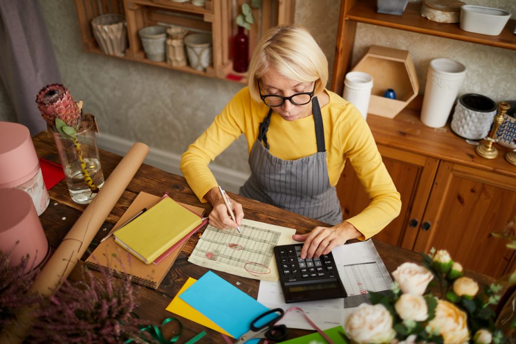 High angle portrait of female businesswoman counting finances using calculator in small shop, copy space. Concept: revise business plan to increase profit