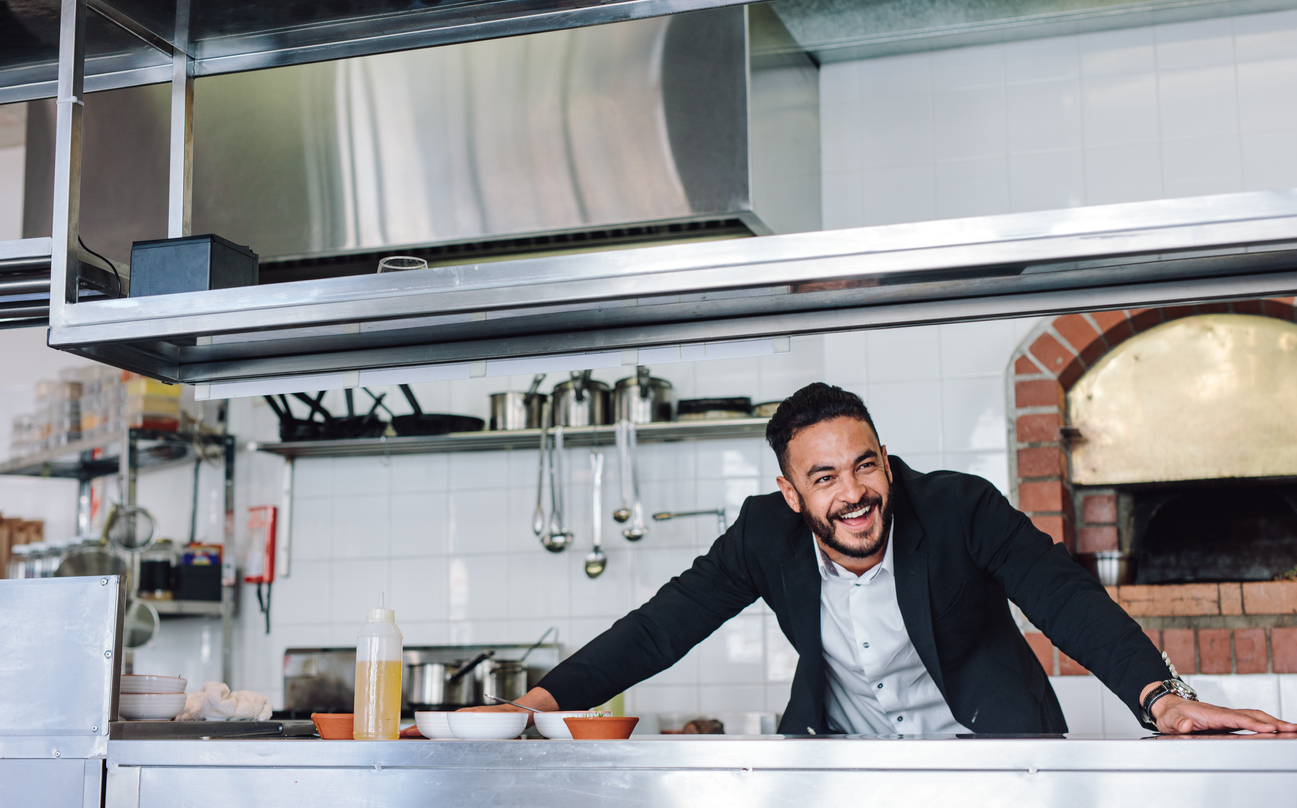 Happy young restaurant owner standing at kitchen counter looking away and smiling. Caucasian businessman in commercial kitchen. Concept: entrepreneur