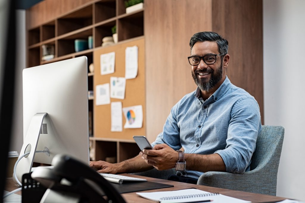 Smiling business man wearing eyeglasses working on desktop computer while using phone in office. Middle eastern businessman using smartphone while sitting at desk in office designer. Man working in modern business environment. Concept: types of loans