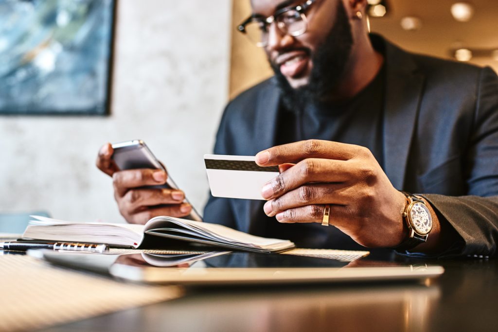 Portrait of African-American male holding cell phone in one hand and unsecured credit card in other, making transaction, using mobile banking app during lunch at cafe. Concept: what is an unsecured credit card?