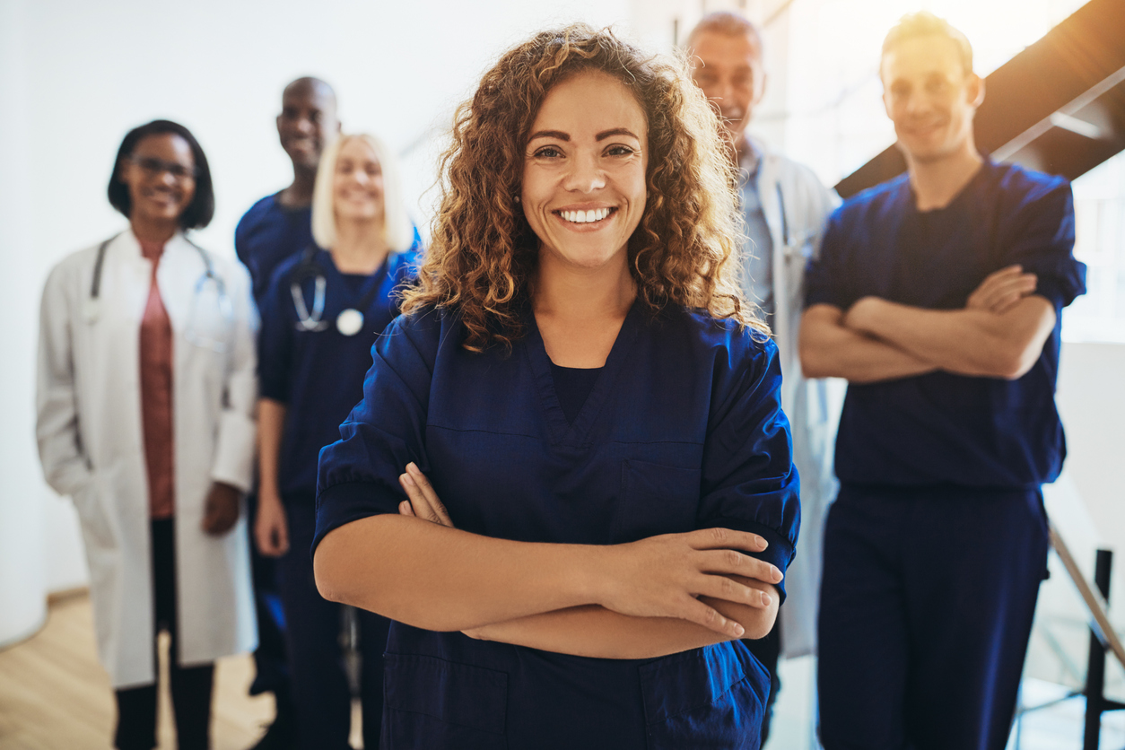 Young female doctor smiling while standing in a hospital corridor with a diverse group of staff in the background. Concept: how to choose health insurance