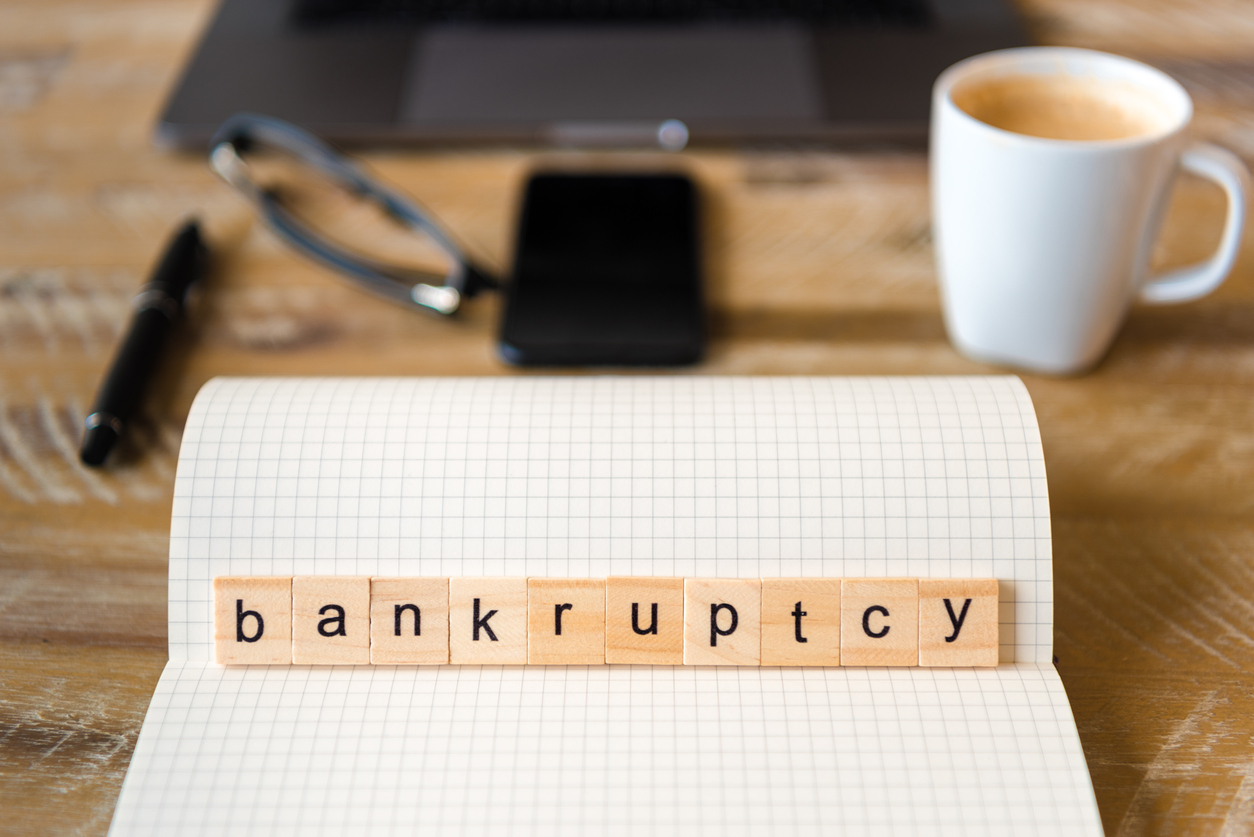 Closeup on notebook over wood table background, focus on wooden blocks with letters making Bankruptcy word. Business concept image. Laptop, glasses, pen and mobile phone in a defocused background. Concept: rebuild credit