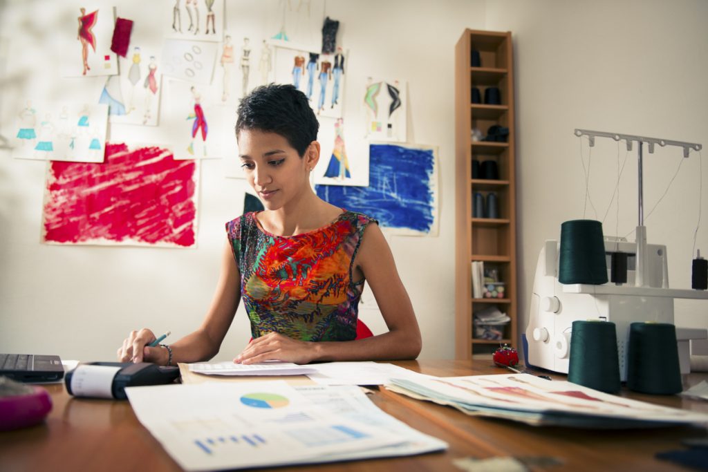Money and financial planning, young hispanic self-employed woman checking bills and doing budget with calculator, computer and papers in fashion design studio. concept: paypal loan