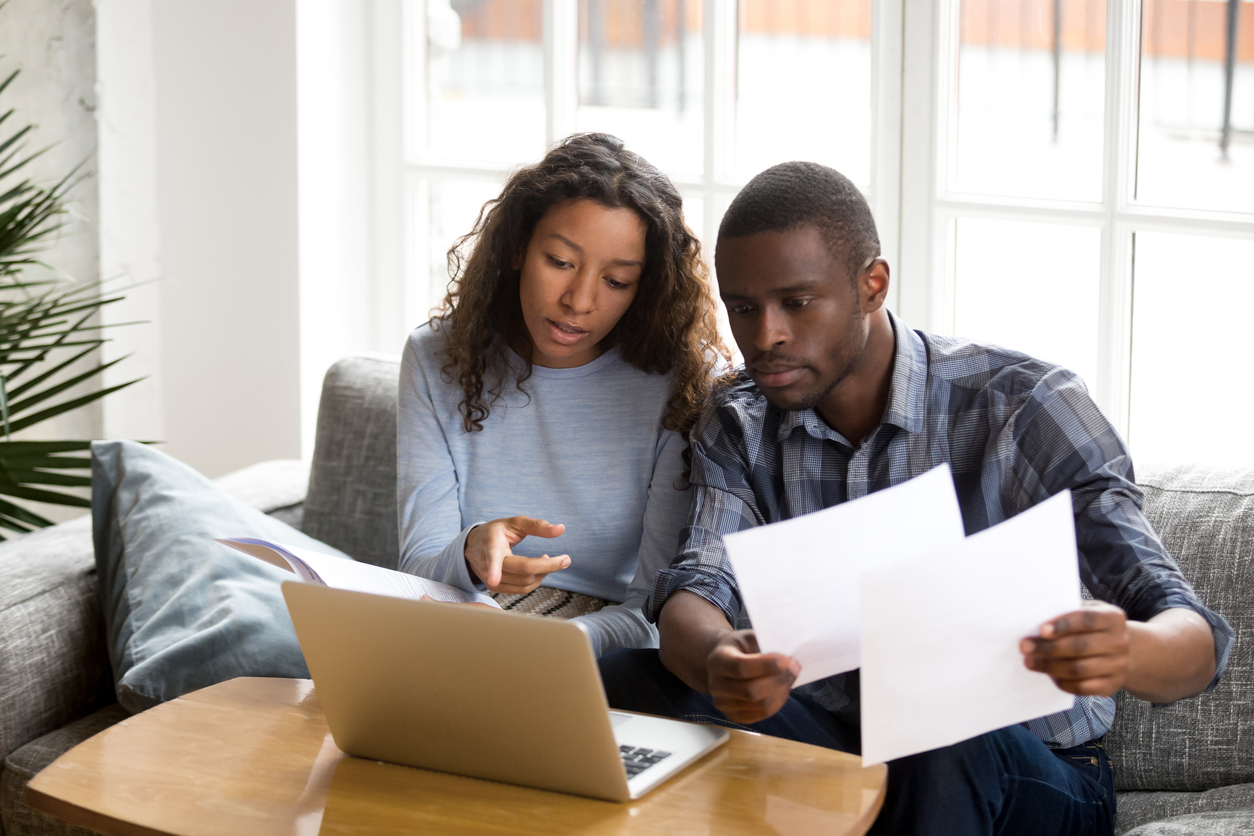 Serious African American couple discussing paper documents, sitting together on couch at home, man and woman checking bills, bank account balance, terms of contract, mortgage, loan agreement. concept: Business Loan Marketplace