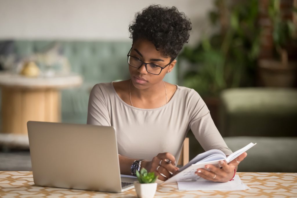 Focused young african american businesswoman or student looking at laptop holding book learning, serious black woman working or studying with computer doing research or preparing for exam online. Concept: Best Entrepreneur Books for Women