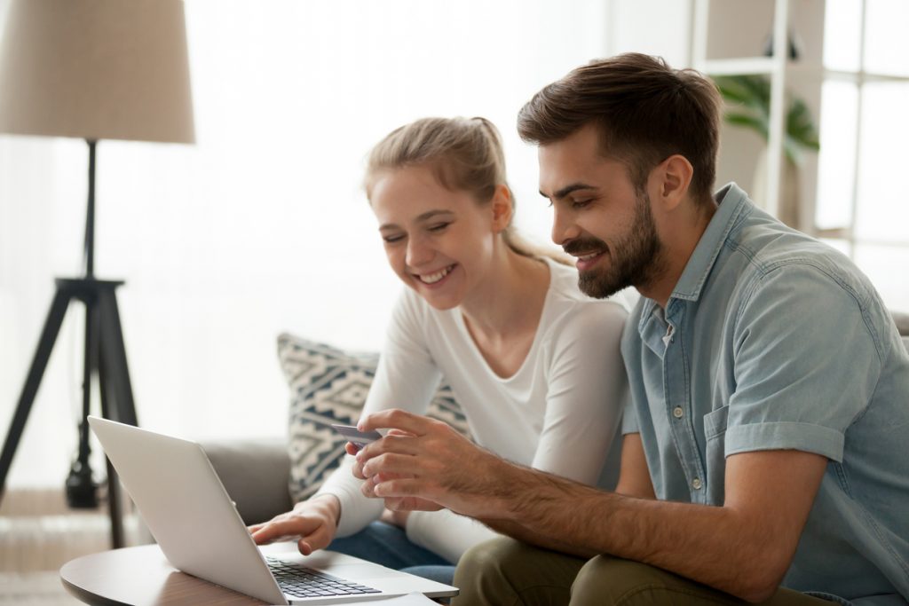 Happy married couple sitting in living room on couch at home, using computer purchasing online. Husband holding credit card customers making secure payment via internet. Technology, e-banking. concept: recurring payments