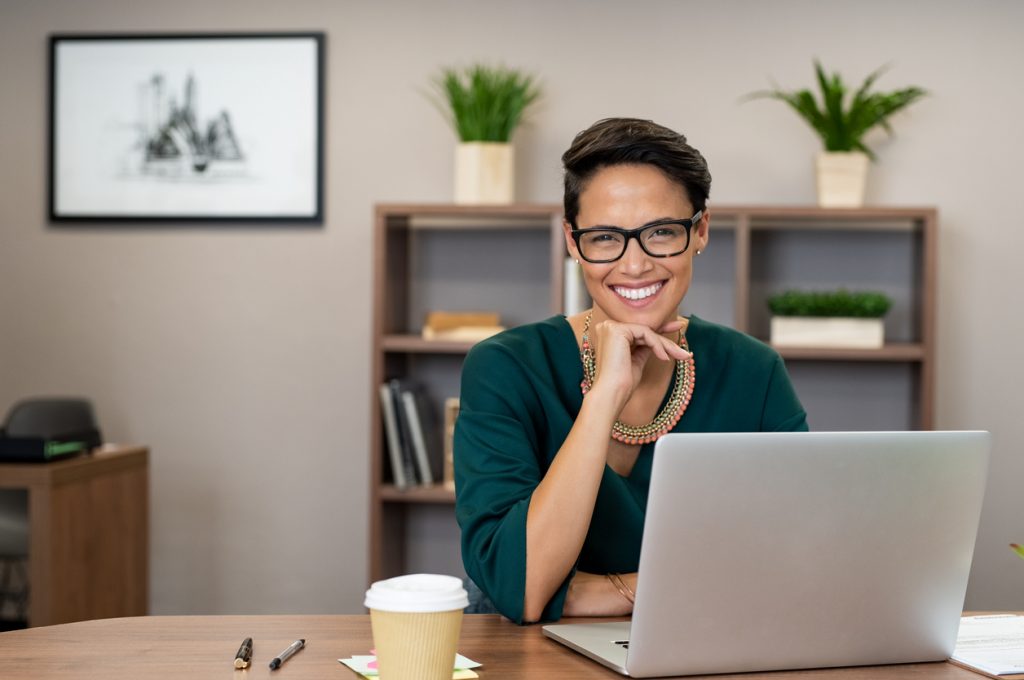 Portrait of happy casual businesswoman sitting at her desk in office. Cheerful latin woman working on laptop and looking at camera. Young fashionable girl wearing eyeglasses sitting at creative agency. Concept: business terms