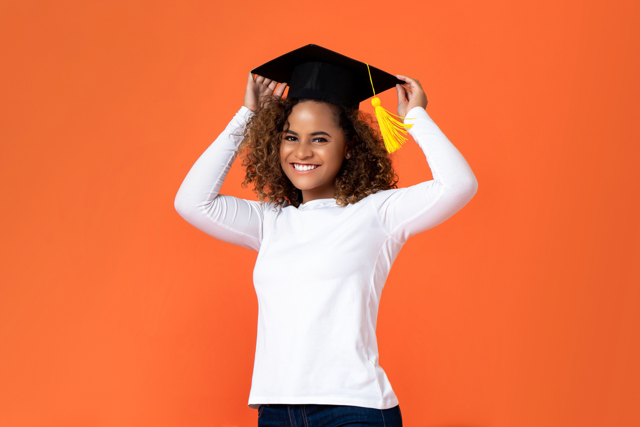 Happy smiling young African American woman wearing graduation cap isolated on orange background for education concepts, camino financial