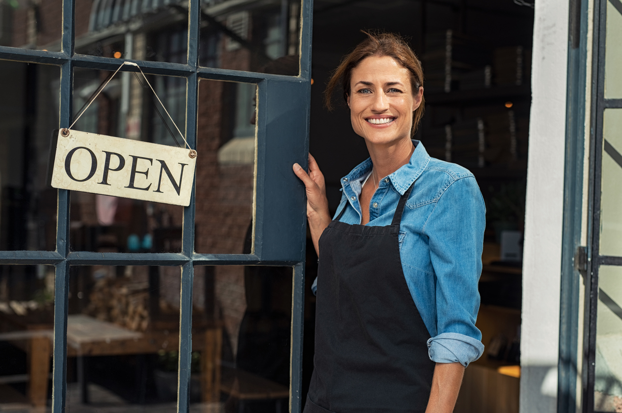 Portrait of a happy waitress standing at restaurant entrance. Portrait of mature business woman in black apron ready to attend new customers in her just opened coffee shop. Happy beautiful woman owner showing open sign in her small business shop. concept: Restaurant reopening plan