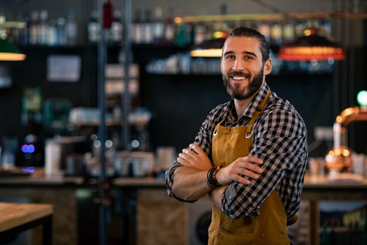 Happy satisfied bartender wearing apron while standing near counter with crossed arms and looking at camera. Confident coffee shop waiter smiling at cafeteria. Successful proud young brewer at his pub with copy space. concept: business mistakes